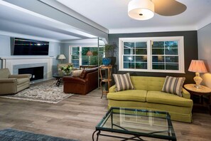 A spacious living room with a fireplace, a green sofa, a glass coffee table, and a flat-screen TV mounted above the fireplace. - Grosse Pointe Windmill Pointe 3BR Upper Flat – Spacious, Airy and Spotless (Grosse Pointe Park)