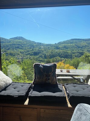 A room with a view of a mountainous landscape, a wooden table with cushions, and a window.