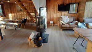 A cozy cabin interior with a central wood stove, a sofa, a dining table, and a TV mounted on the wall.