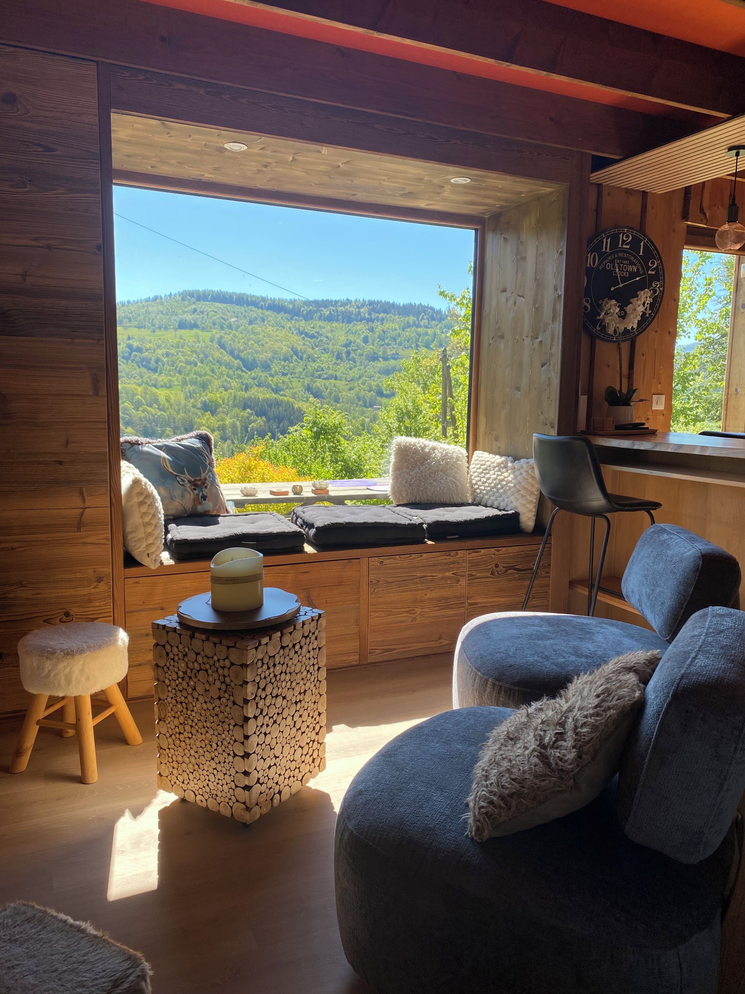 A cozy room with a large window, wooden furniture, and a view of a forested hillside.