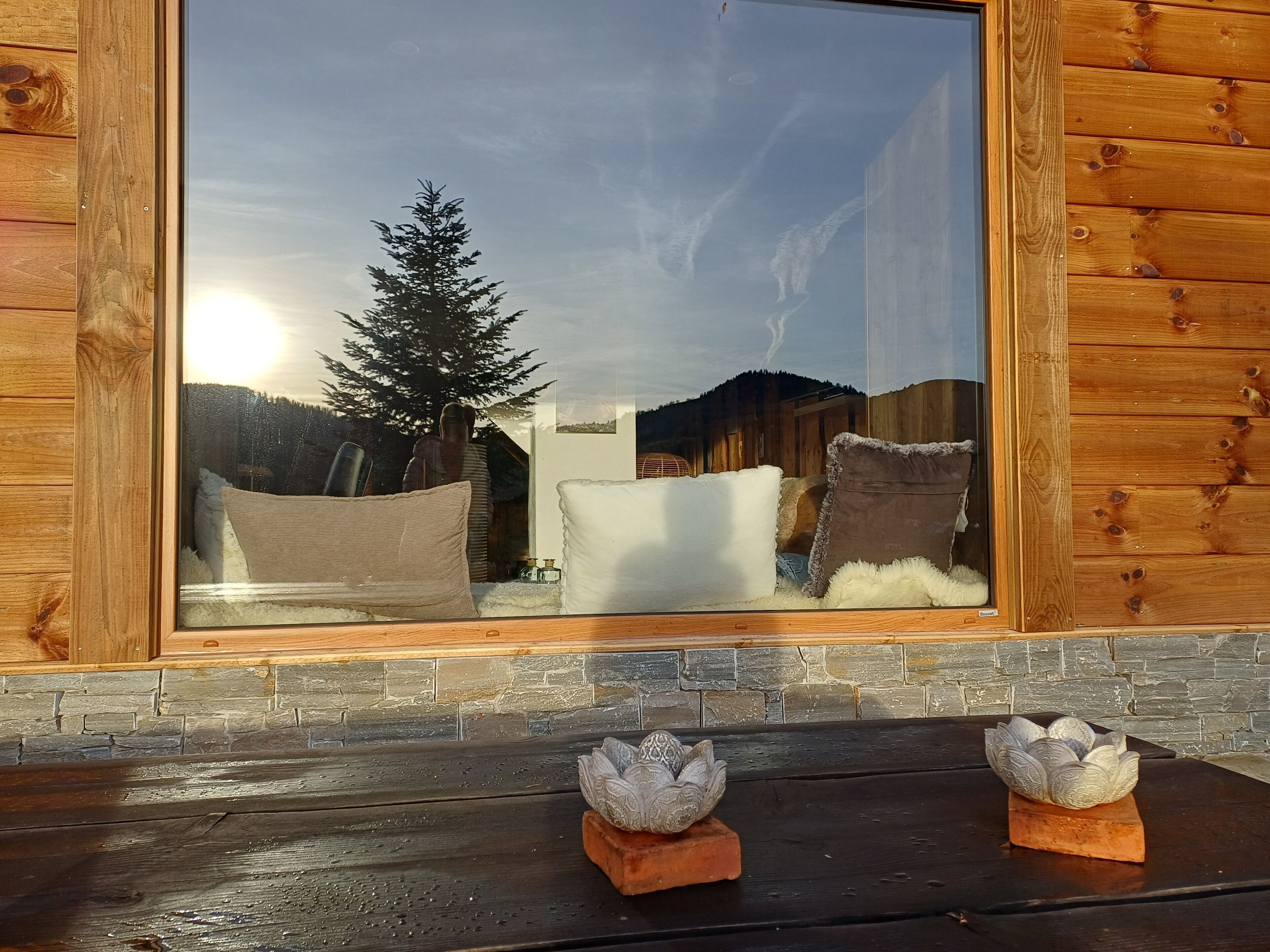 Une cabane en bois dotée d’une grande fenêtre qui reflète un paysage enneigé, d’une cheminée en pierre et de deux vases décoratifs en forme de lotus, placés sur le manteau de la cheminée.