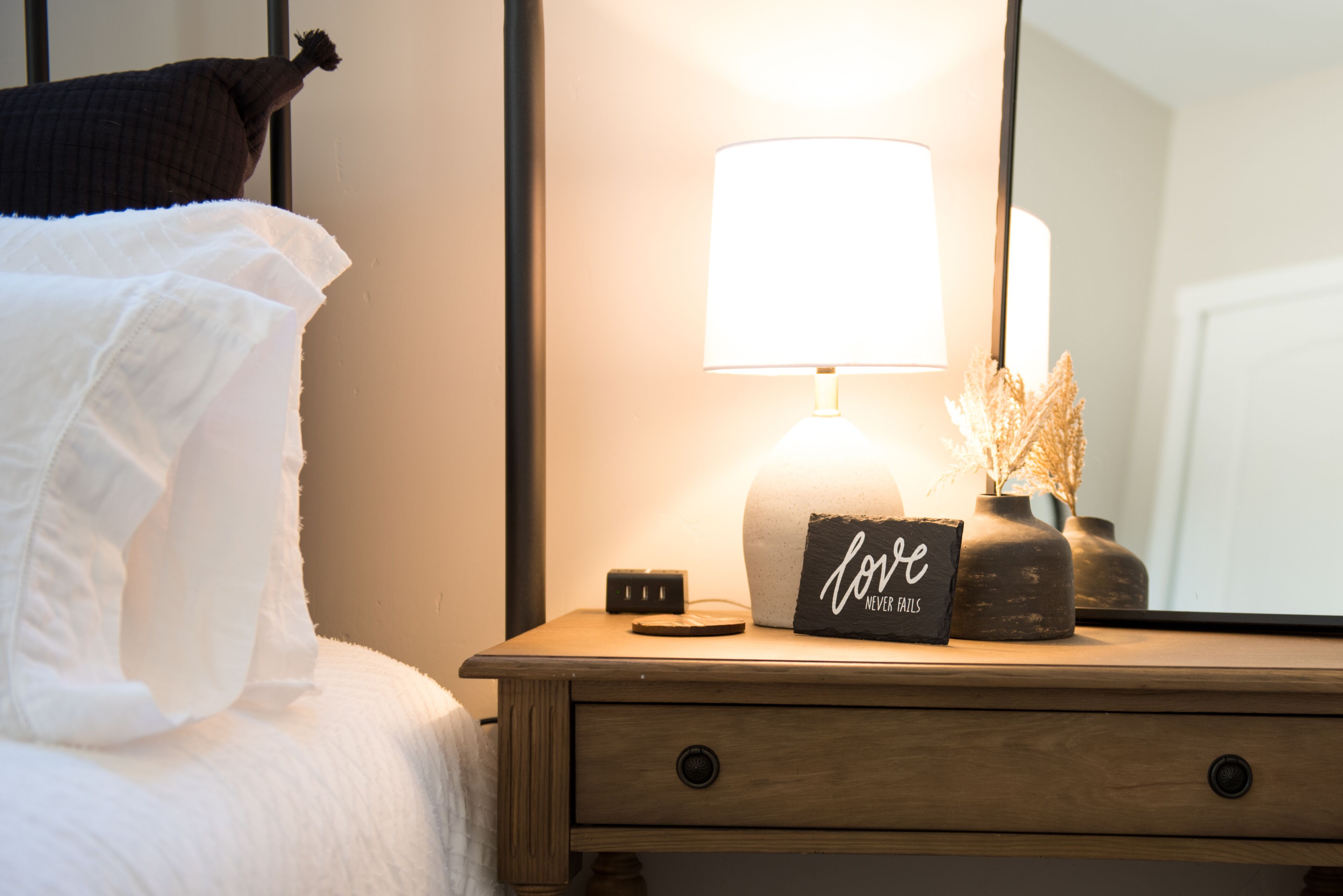 Una cama bien hecha con sábanas blancas, una lámpara de noche, un pequeño reloj y un cartel que dice "Love Never Fails" sobre una mesita de noche de madera.