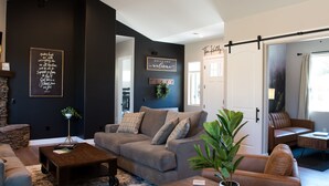 A modern living room with a grey sofa, a coffee table, and a potted plant. There is a sign on the wall and a door leading to another room. - Custom 4bd Mountain & Golf Retreat, Sleeps 8,
A/C, Close to All Key Attractions (Williams)
