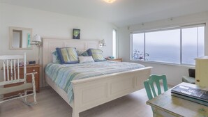 A bedroom with a bed, a chair, a table, a nightstand, a lamp, a framed picture, and a window with a view.