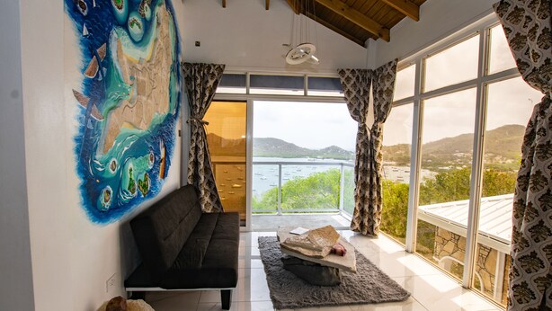 Een ruime kamer met een groot raam, met uitzicht op zee en heuvels, een zwarte bank, een vloerkleed en een schilderij aan de muur.