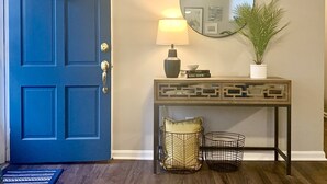 A blue door with a wreath, a lamp, a mirror, a potted plant, and a console table with decorative items.