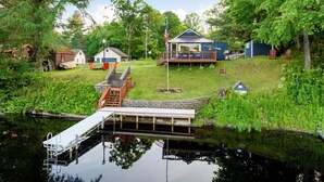 En stuga vid sjön med en brygga, en liten veranda och en flaggstång.