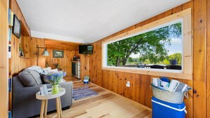 En mysig stuga med träpanel på insidan, en soffa, ett litet bord med en vas, ett fönster med utsikt över träd och en blå kylbox.