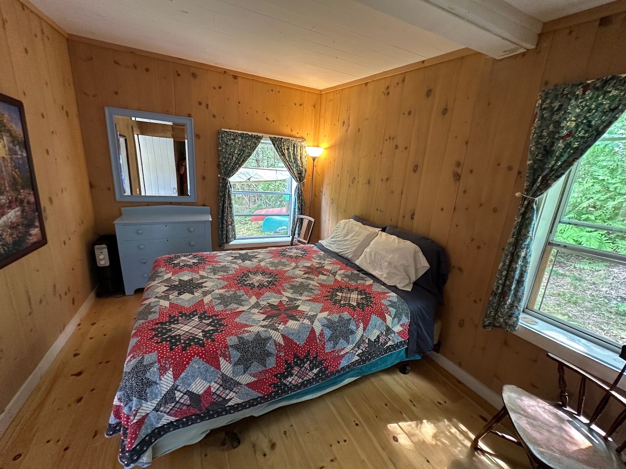 Un dormitorio con piso de madera, una cama con colcha estampada, un aparador, un espejo y una ventana con cortinas.
