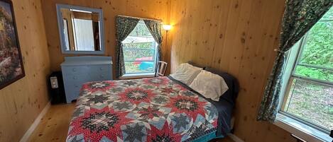 Un dormitorio con piso de madera, una cama con colcha estampada, un aparador, un espejo y una ventana con cortinas.