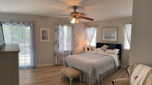 Quarto com cama, mesa de cabeceira, cadeira, banqueta, ventoinha no teto e cortinas.