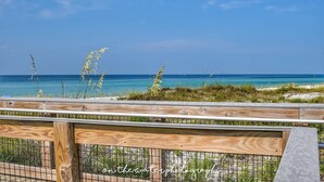 A wooden deck with a metal railing overlooking a beach with clear blue water and sparse vegetation. - Quiet Family Friendly Saltwater Pool Home, Near Beach (Navarre)