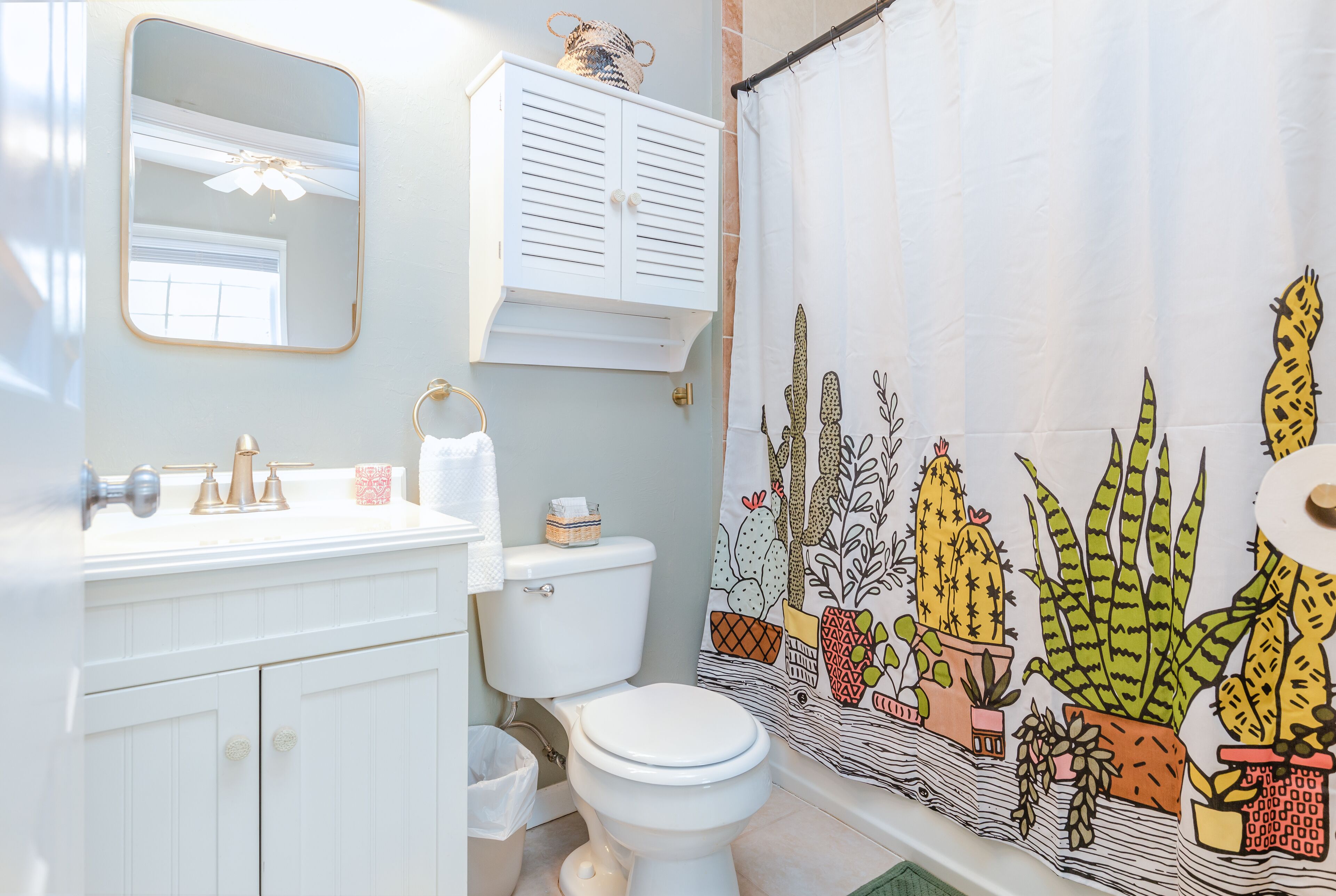 Ein Badezimmer mit Toilette, Waschbecken, Spiegel, Schrank und Duschvorhang mit Kaktusmuster.