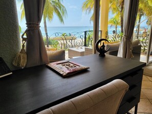 Quarto com escrivaninha de madeira escura, uma cadeira e vista para o oceano através de grandes janelas.