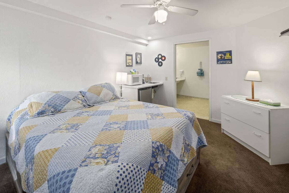 Een slaapkamer met een bed, een bureau, een stoel, een lamp en een plafondventilator.