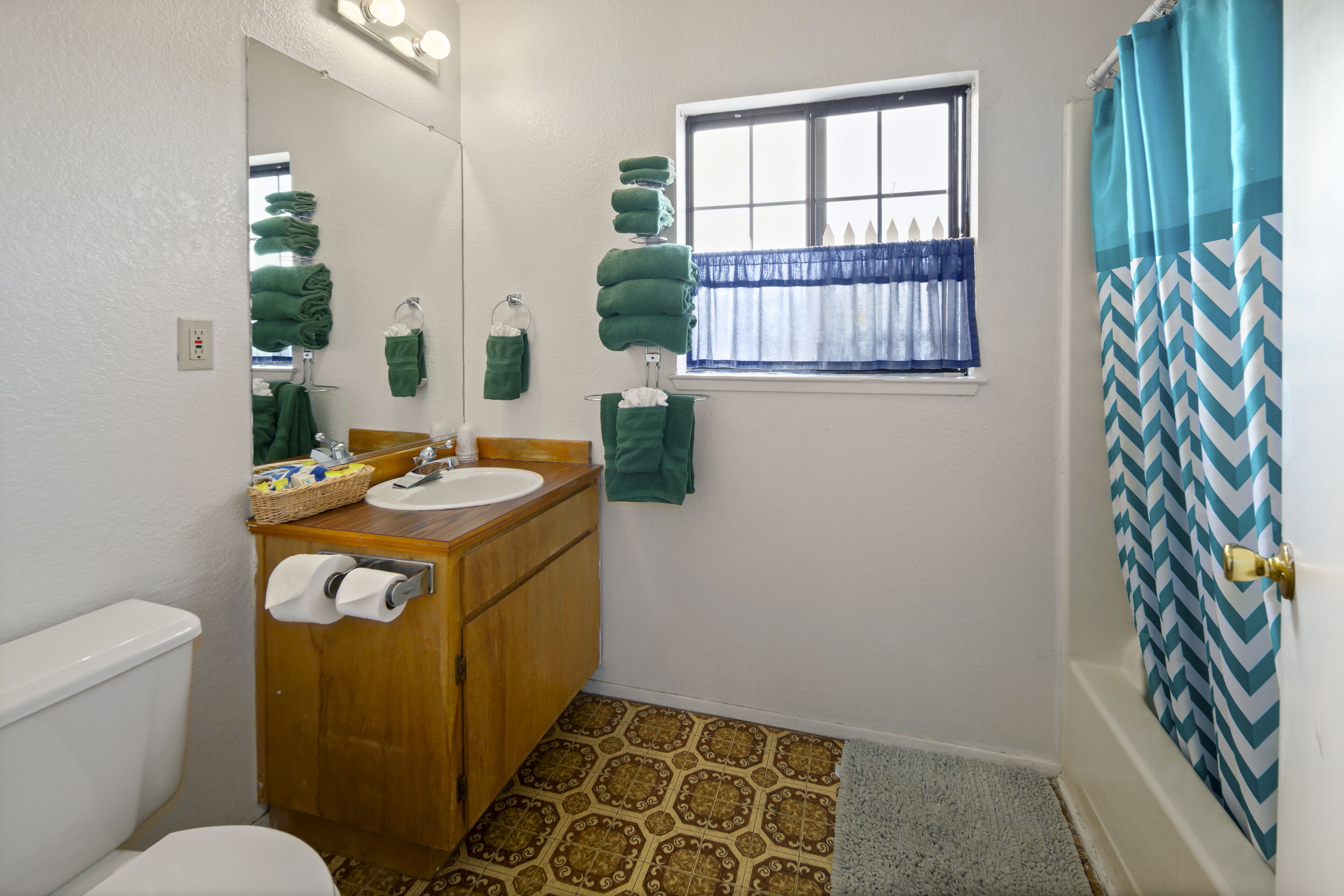 Een badkamer met toilet, wastafel en een wastafelmeubel. Er is een raam met een blauw gordijn en een douche met een ruitpatroon gordijn.