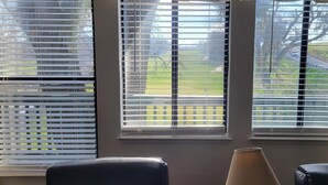 Una habitación con vistas a través de una ventana con persianas, una lámpara y dos sillas.