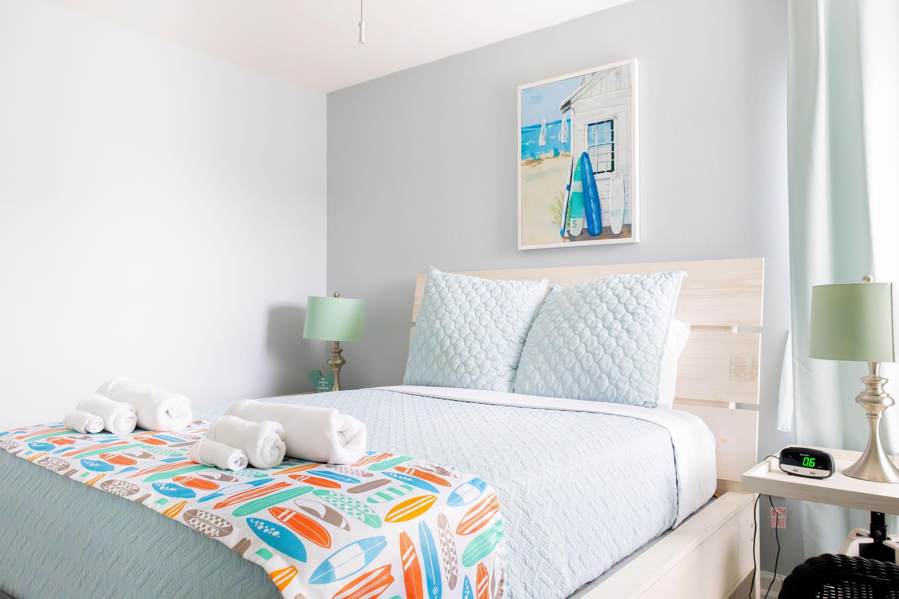 A neatly made bed with a quilted headboard, a bedside table with a lamp, and a wall-mounted painting of a beach scene.