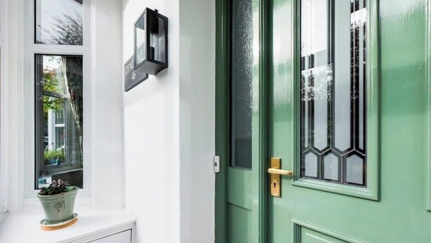 Eine grüne Tür mit Fenster, ein Blumentopf auf einem weißen Schrank und eine schwarze Wandleuchte.