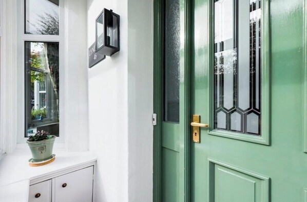 A green door with a window, a potted plant on a white cabinet, and a black wall-mounted light fixture. - Windermere Cottage for 8 | Walk Everywhere. Authentic Lakeland slate cottage, (Windermere)
