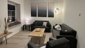 A modern living room with a sofa, a coffee table, and a bench. There is a window with blinds and a lamp on a side table. - Vacation home "de Fofftein" in Bockhöft (Ellingstedt)