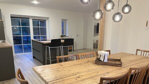 A modern kitchen with a central island, a dining area with a wooden table and chairs, and a view of the backyard through large windows. - Vacation home "de Fofftein" in Bockhöft (Ellingstedt)