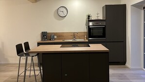 A modern kitchen with a wooden island, black cabinets, a built-in oven, and a wall-mounted clock. - Vacation home "de Fofftein" in Bockhöft (Ellingstedt)