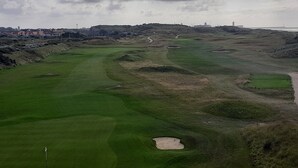 A golf course with bunkers, a flagstick, and a distant view of buildings and a lighthouse.