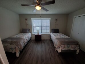 Quarto com duas camas, um ventilador de teto, uma janela com persianas e uma mesa de cabeceira com abajur.