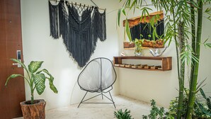 A room with a macrame wall hanging, a chair, a shelf with books, and various plants. - Moonshine Tulum Hotel  (Tulum)