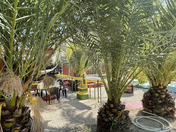 A covered outdoor area with palm trees, a yellow and green bench, and a table with chairs.