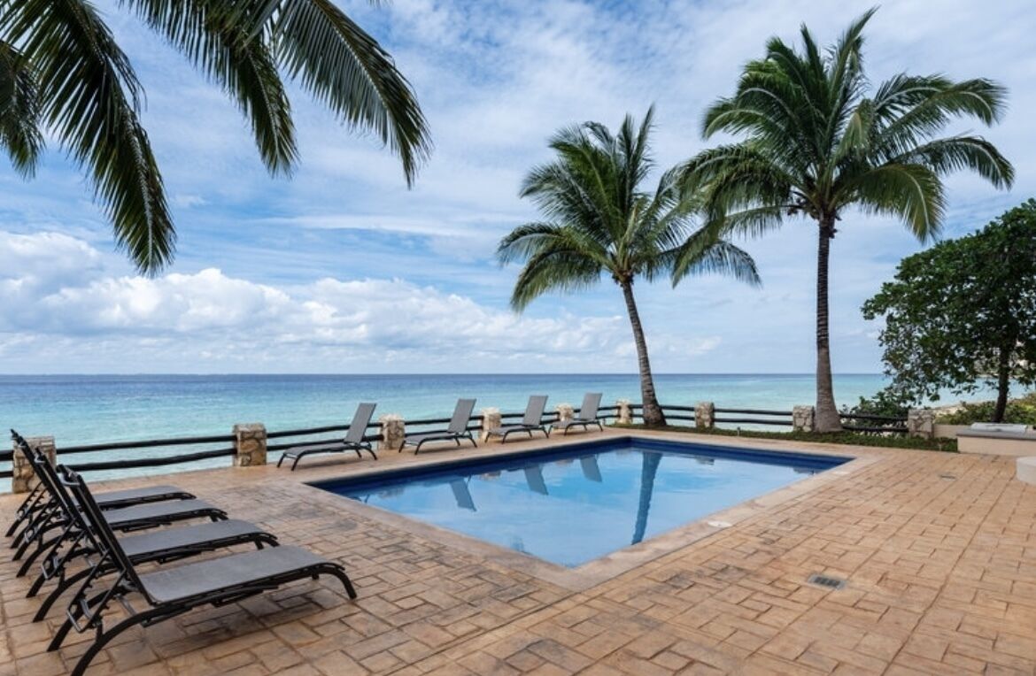 A swimming pool with lounge chairs, palm trees, and a view of the ocean.