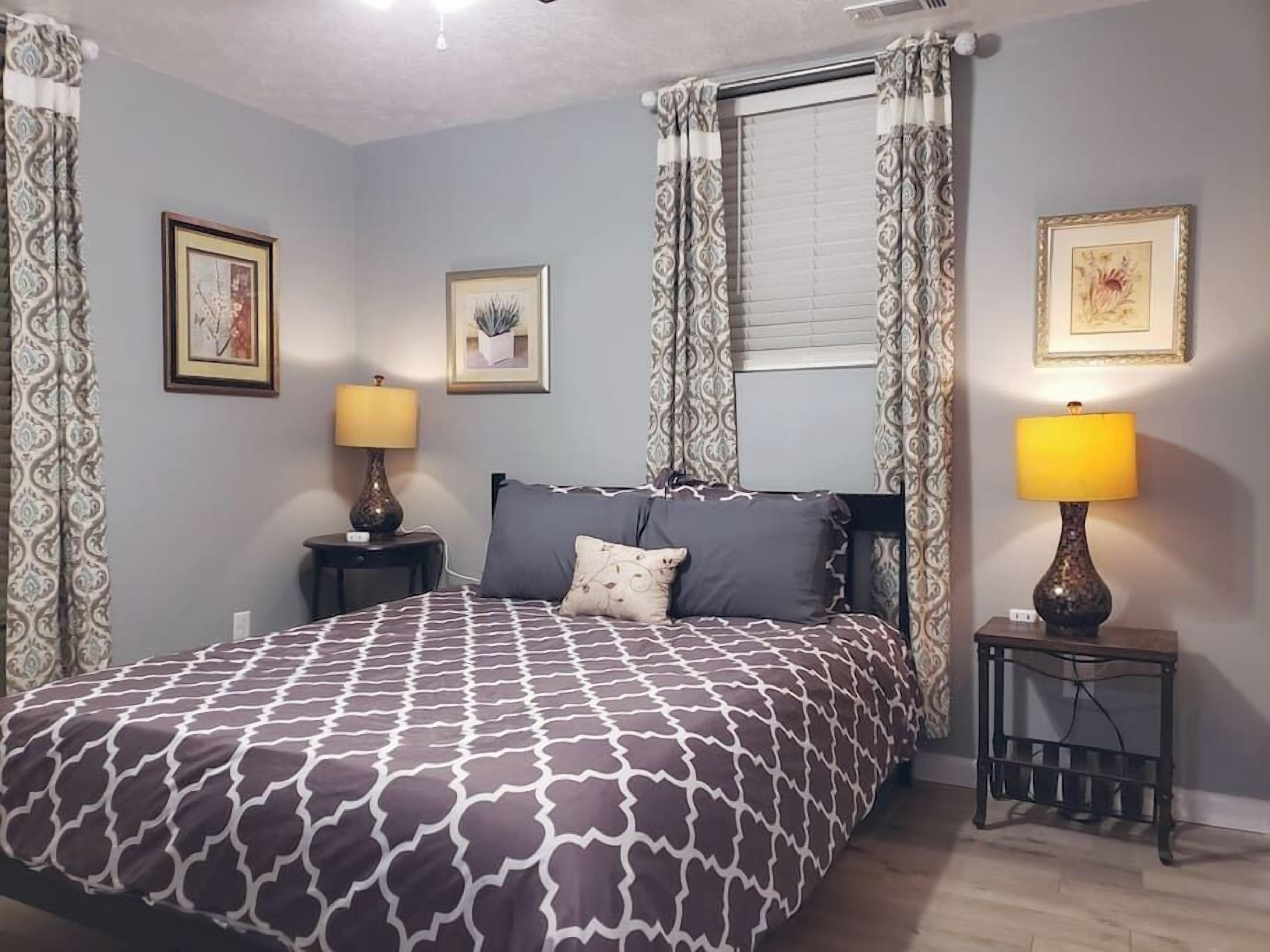 A bedroom with a patterned bedspread, two bedside lamps, a nightstand, and two framed pictures on the wall.