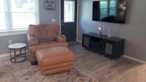 Un salon avec un fauteuil en cuir, un pouf et un téléviseur à écran plat posé sur un meuble noir.