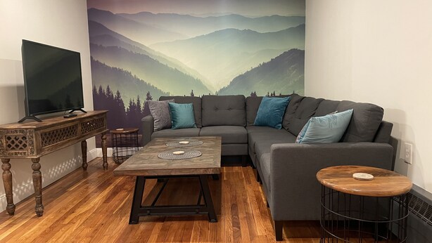 A living room with a grey sofa, a coffee table, a TV on a wooden stand, and a mural of mountains. - Newly renovated1 Bedroom 1 Bath in the Heart of Downtown Ellsworth.  (Ellsworth)