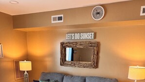 Un salon avec un canapé d’angle gris, une table basse blanche et deux petites tables d’appoint avec des lampes. Un miroir avec un cadre rustique et une pancarte sur laquelle on peut lire « ADMIRONS LE COUCHER DE SOLEIL » sont accrochés au mur.