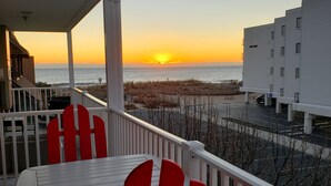 Un balcon offrant une vue sur le coucher de soleil sur l’océan, un bâtiment blanc en arrière-plan et des chaises rouges.