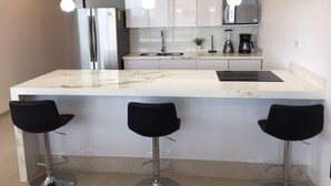 A modern kitchen with a marble countertop, stainless steel appliances, and three black bar stools. - OCEAN VIEW APARTMENT (Dzemul,)