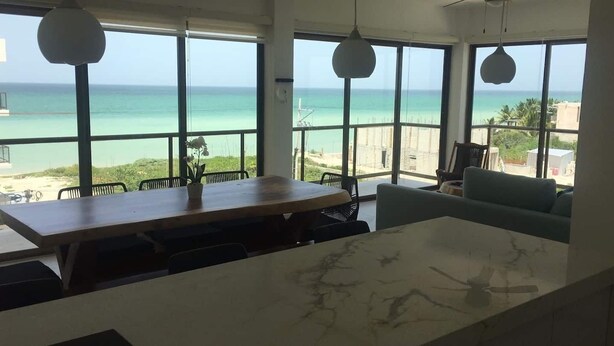 A room with a large window overlooking a beach, a wooden table, a marble countertop, and a green sofa. - OCEAN VIEW APARTMENT (Dzemul,)