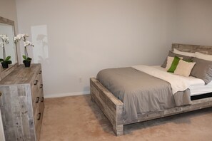 Ein Schlafzimmer mit einem Holzbettgestell, beigefarbener Bettwäsche und einer Kommode mit Orchideen in Töpfen.