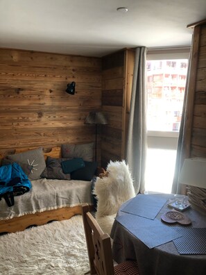 A cozy room with wooden walls, a sofa with cushions, a fluffy white rug, a small round table with a lamp, and a window with curtains. - Tignes Val Claret: cocoon at the foot of the slopes (Tignes Val Claret)