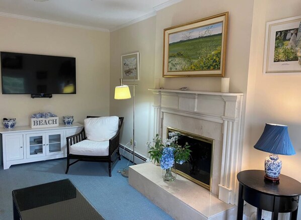 A living room with a fireplace, a television, a white armchair, a blue vase with flowers, and a lamp on a side table.