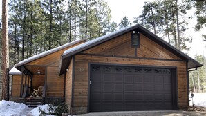 A wooden cabin with a garage, surrounded by snow and trees. - Cozy mountain retreat at 7,200 feet. - Sleeps 9 (Pinetop)
