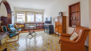 A living room with a sofa, chairs, a television, a cabinet, and a window with a view of buildings outside. - 3 bedroom pet friendly home in Piombino (Piombino)
