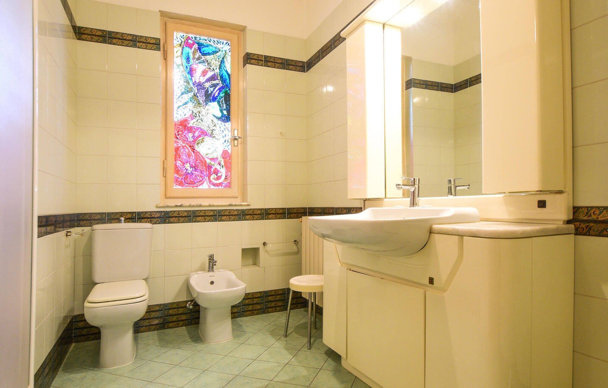 Een badkamer met toilet, bidet, wastafel en een glas-in-loodraam.