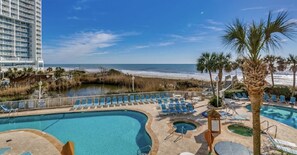 A resort pool area with a view of the beach and ocean, including a large pool, lounge chairs, and palm trees.