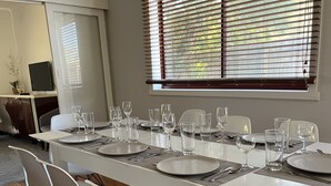 Zona pranzo con un tavolo lungo apparecchiato per sei persone, sedie bianche e una finestra con tende.