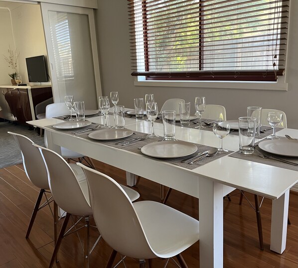A dining area with a long table set for six, white chairs, and a window with blinds.