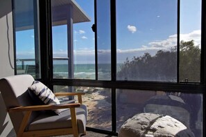 A room with a large window overlooking a beach and ocean, a wooden chair, and a small ottoman. - Beachfront ocean-view apartment with balcony (Semaphore Park)