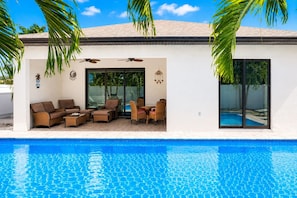 Un área junto a la piscina con un edificio blanco, mobiliario exterior y una piscina azul cristalina.